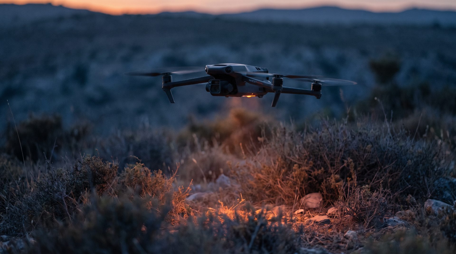 Thermal-equipped quadcopter hovering low over dry scrubland at dusk